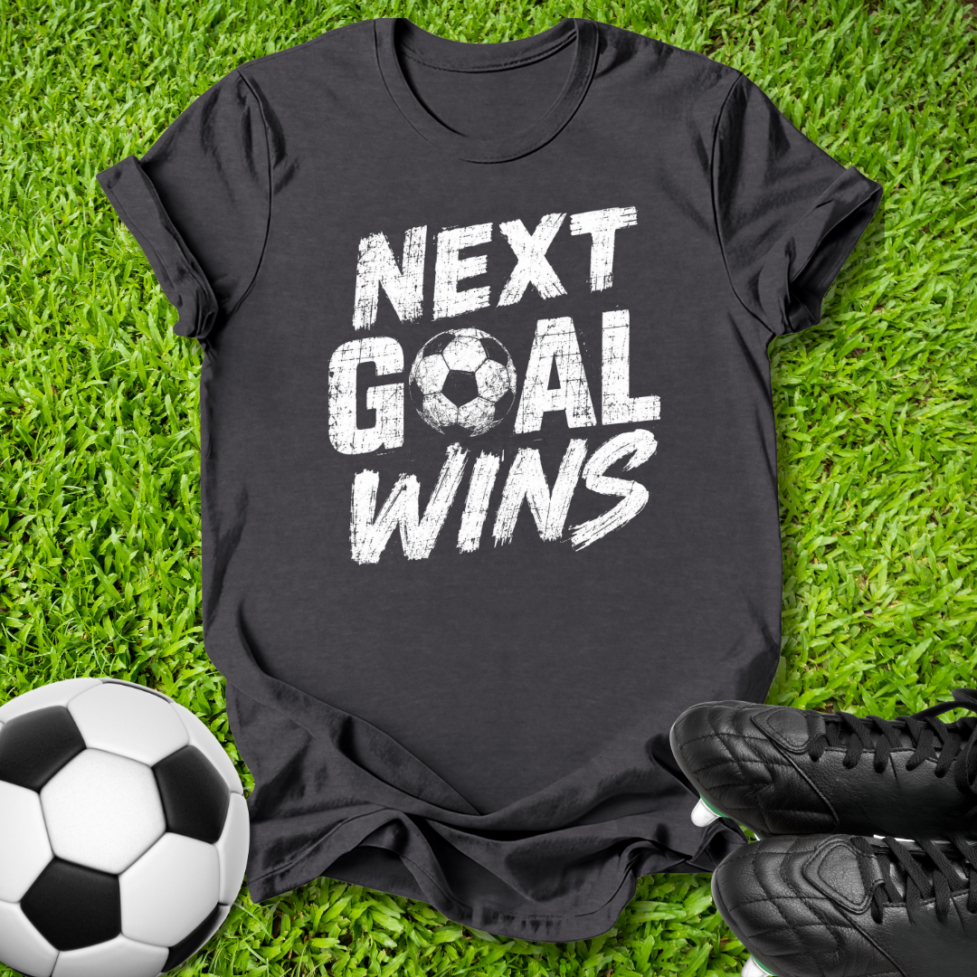 A black short-sleeve graphic t-shirt laid flat on green grass, featuring the phrase 'NEXT GOAL WINS' in distressed white lettering with a soccer ball icon. A black-and-white soccer ball is on the left, and black cleats are on the right.
