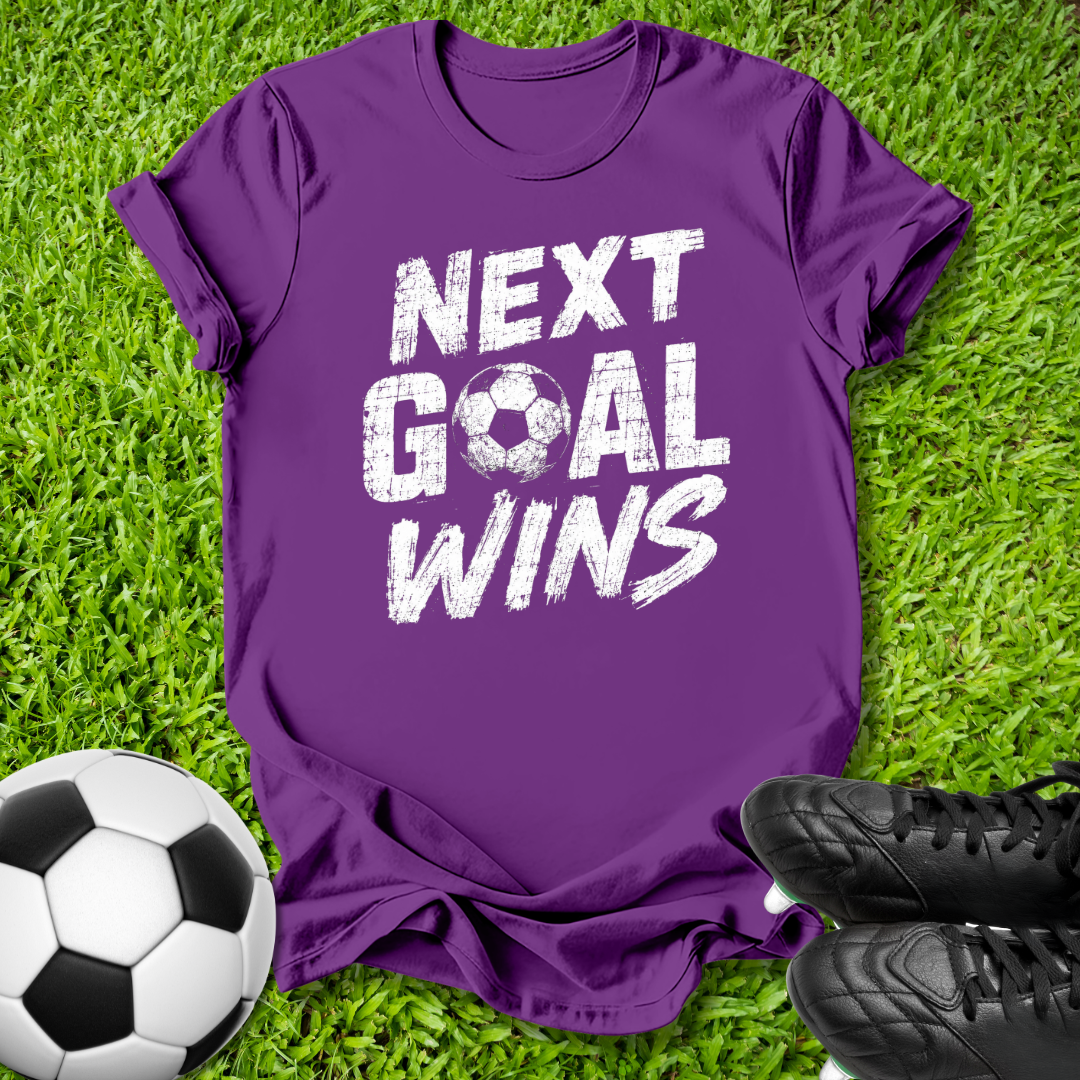 A purple short-sleeve t-shirt with the text 'NEXT GOAL WINS' in white distressed lettering, laid flat on green grass with a soccer ball and black cleats as props.