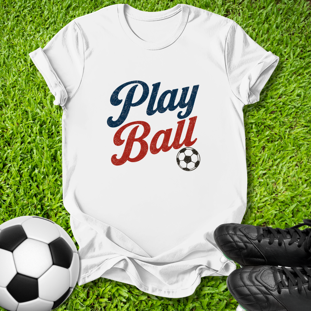 A white short-sleeve T-shirt laid flat on green grass, printed with the words 'Play Ball' in navy and red script and a small soccer ball graphic. A real soccer ball and black cleats are placed around it as props.