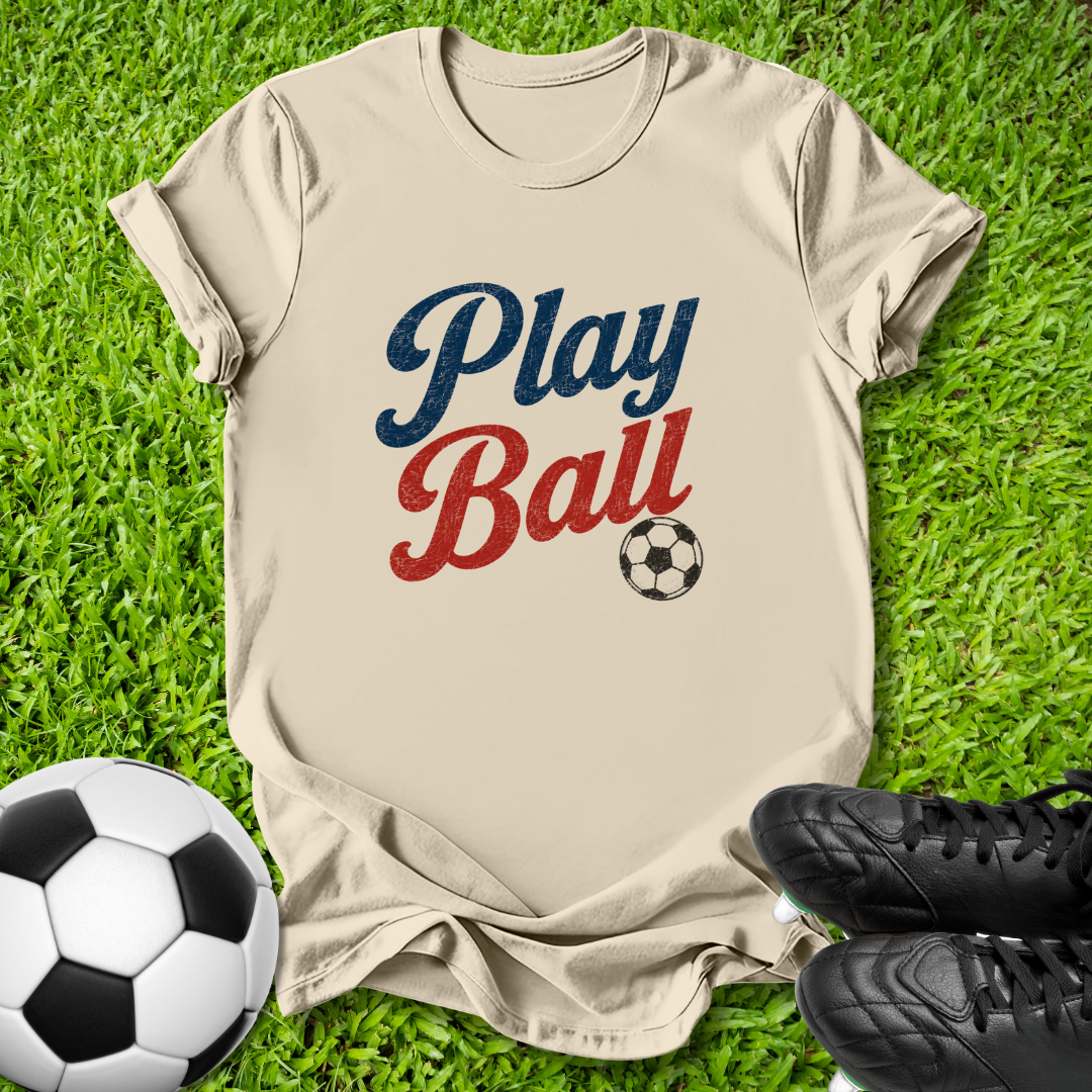 A beige short-sleeve t-shirt with a 'Play Ball' graphic in navy blue and red, styled on green grass with soccer balls and black cleats.