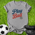 A heather gray t-shirt with a 'Play Ball' graphic in blue and red lettering, laid flat on green grass with soccer balls and black cleats as props.