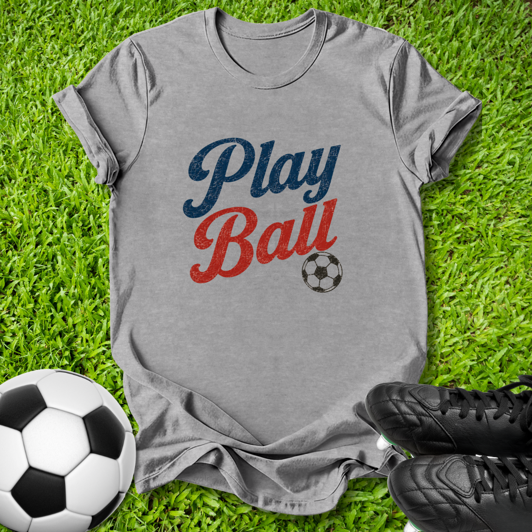A heather gray t-shirt with a 'Play Ball' graphic in blue and red lettering, laid flat on green grass with soccer balls and black cleats as props.