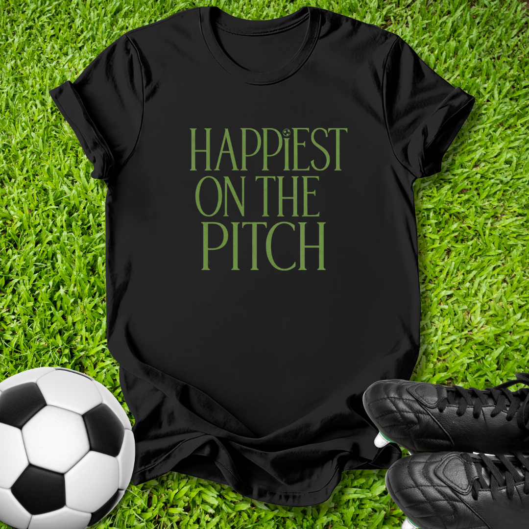 A black short-sleeve t-shirt with the phrase 'HAPPIEST ON THE PITCH' printed in green serif letters, laid flat on green grass with a soccer ball and black cleats as props.