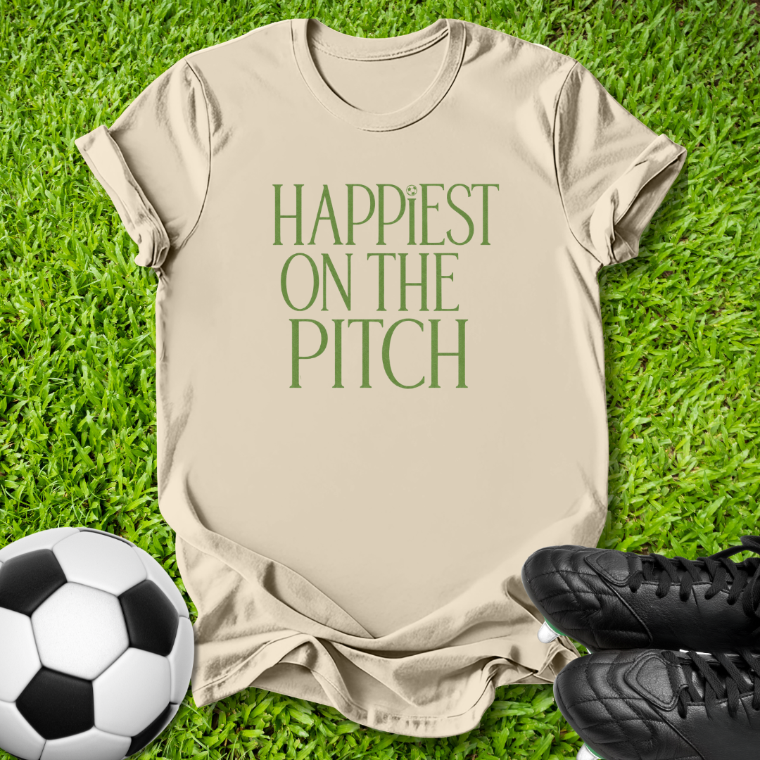 A beige short-sleeve t-shirt with the phrase 'HAPPIEST ON THE PITCH' printed in green, laid flat on green grass with a soccer ball and black cleats as props.