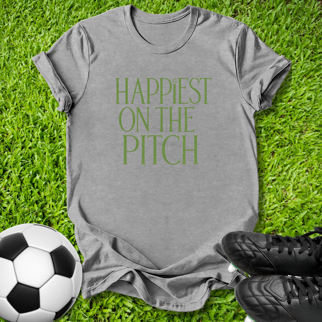 A gray short-sleeve t-shirt with the phrase 'HAPPIEST ON THE PITCH' printed in green serif letters, laid flat on green grass with a soccer ball and black cleats as props.