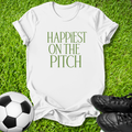 A white short-sleeve T-shirt laid flat on green grass, printed with the phrase 'HAPPIEST ON THE PITCH' in green serif lettering. A soccer ball appears at the lower left, and black soccer cleats are at the lower right.