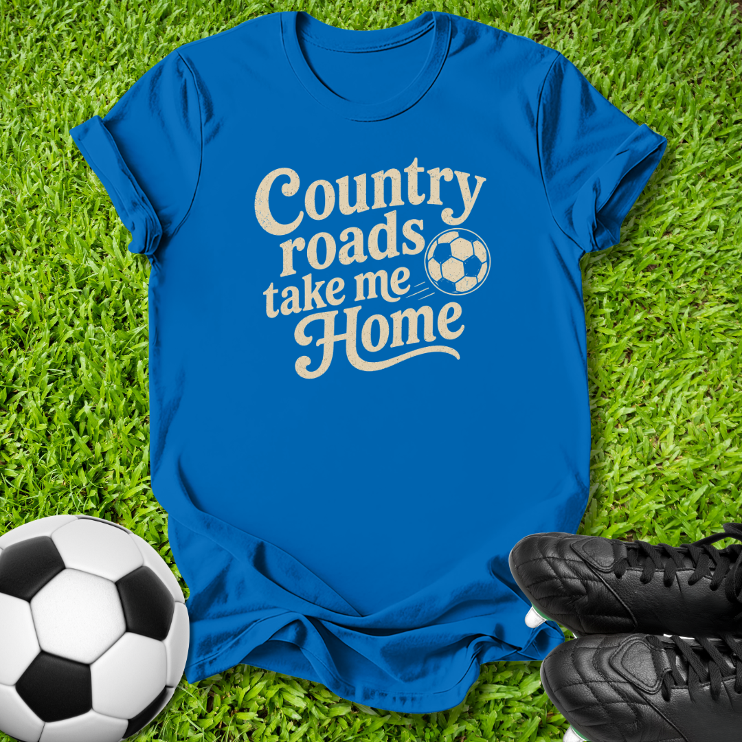 A royal blue short-sleeve t-shirt with the phrase 'Country roads take me Home' in white script, laid flat on green grass with a soccer ball and black cleats nearby.