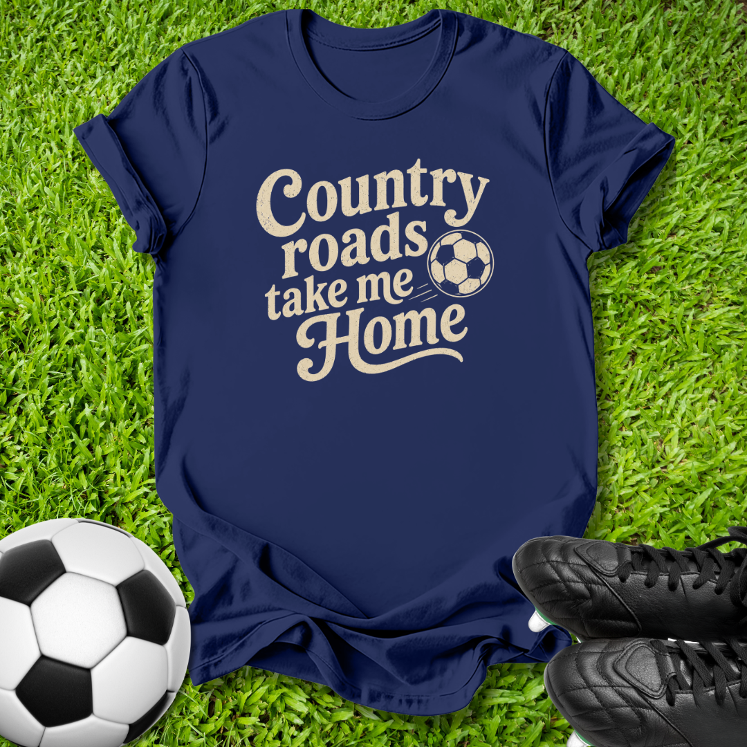 A navy blue short-sleeve t-shirt with the phrase 'Country roads take me Home' in a cream-colored script font, accompanied by a soccer ball graphic. The shirt is laid flat on green grass with a soccer ball and black cleats as props.
