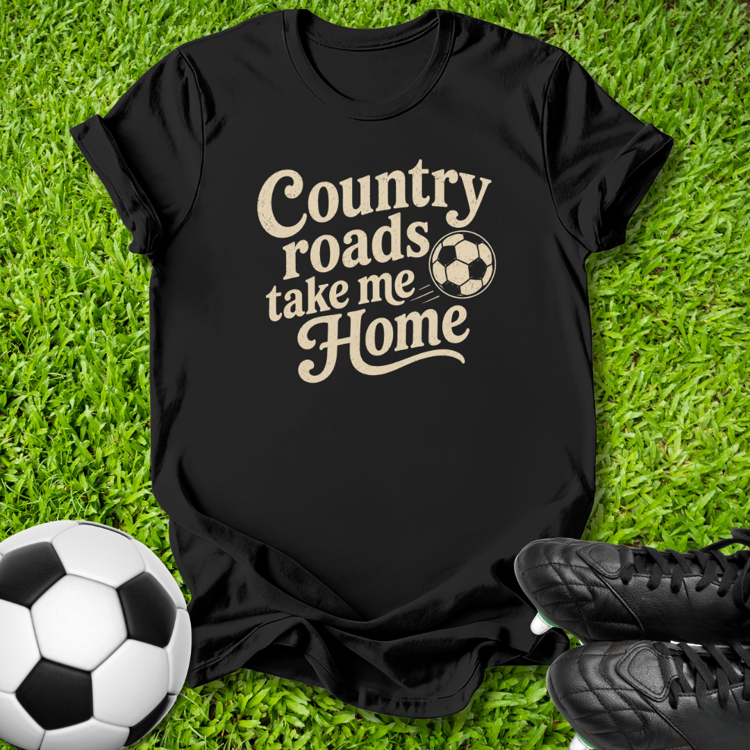 A black short-sleeve graphic T-shirt laid flat on green grass, with the phrase 'Country roads take me Home' in beige lettering and a soccer ball motif. A soccer ball and black cleats are placed around it as styling props.