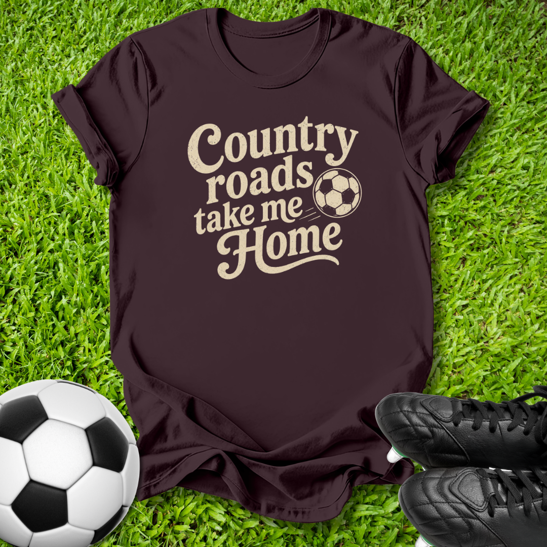 A dark maroon short-sleeve t-shirt with a beige graphic reading 'Country roads take me Home' and a soccer ball motif, laid flat on green grass with a soccer ball and black cleats nearby.