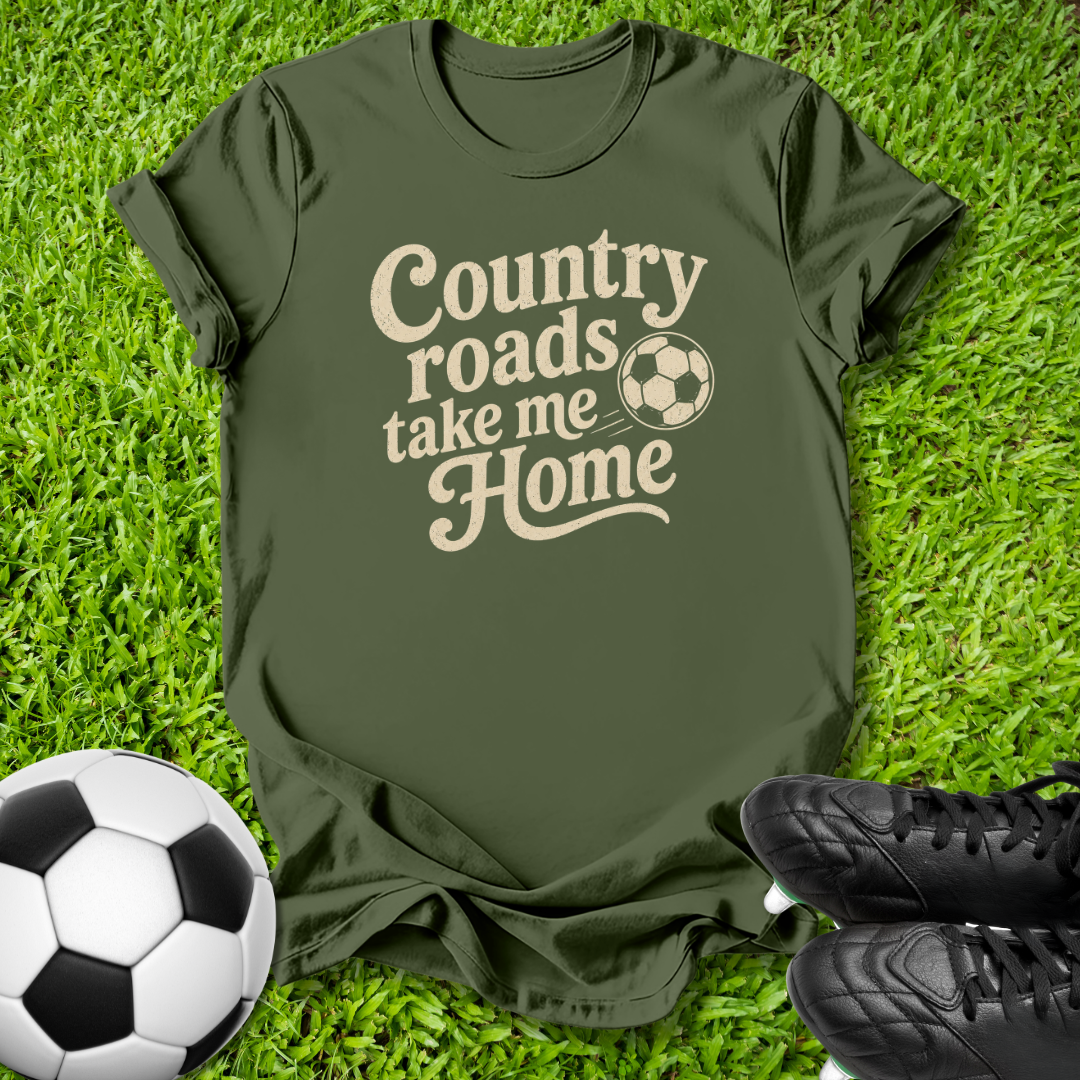 An olive green t-shirt with a beige graphic reading 'Country roads take me Home' and a soccer ball icon, laid flat on green grass with soccer cleats and a soccer ball as props.