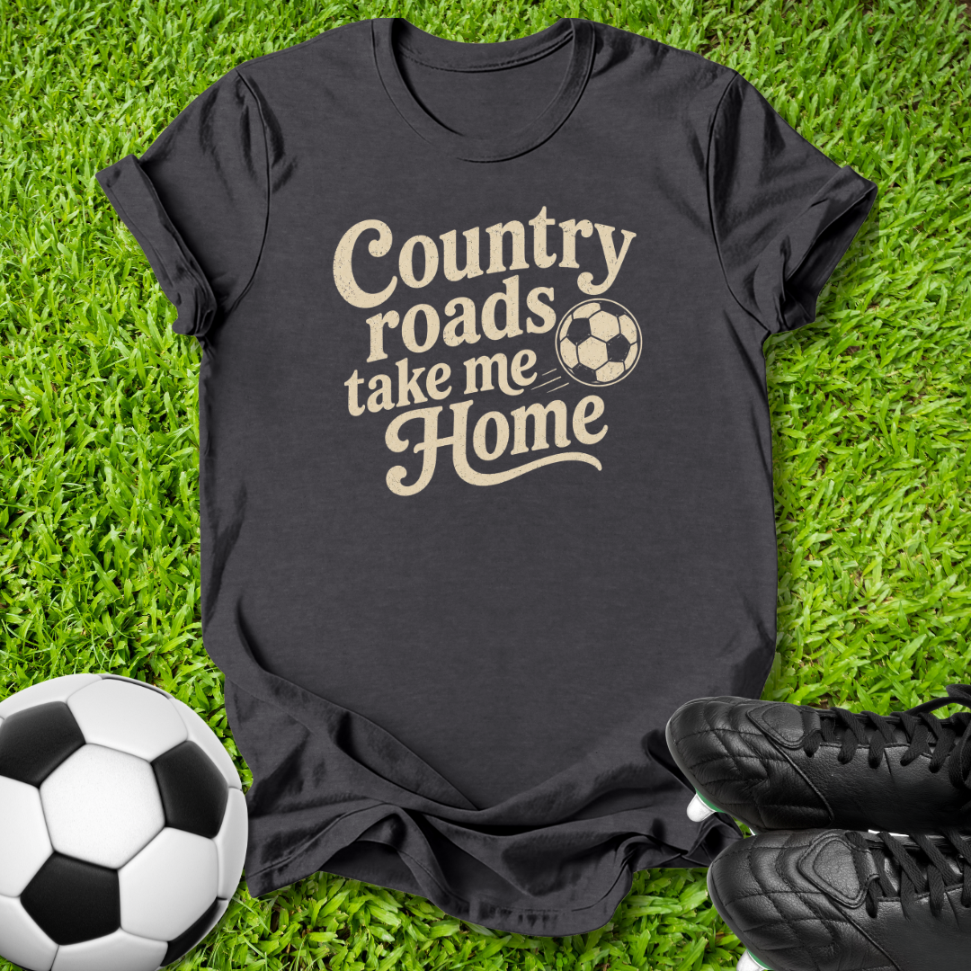A black short-sleeve graphic T-shirt laid flat on green grass, with a beige slogan reading 'Country roads take me Home' and a soccer ball motif. A soccer ball and black cleats are placed around it as styling props.