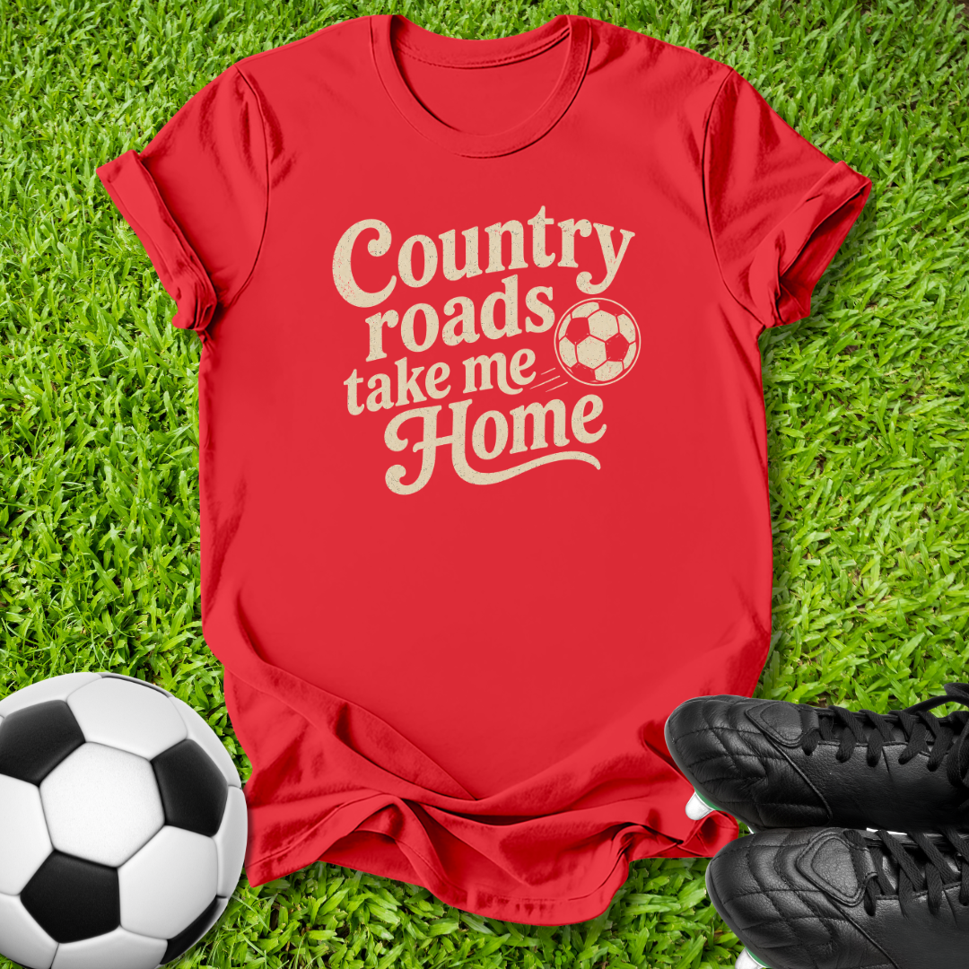 A red short-sleeve t-shirt with the phrase 'Country roads take me Home' in a cream-colored retro font, laid flat on green grass with soccer-themed props including a soccer ball and black cleats.