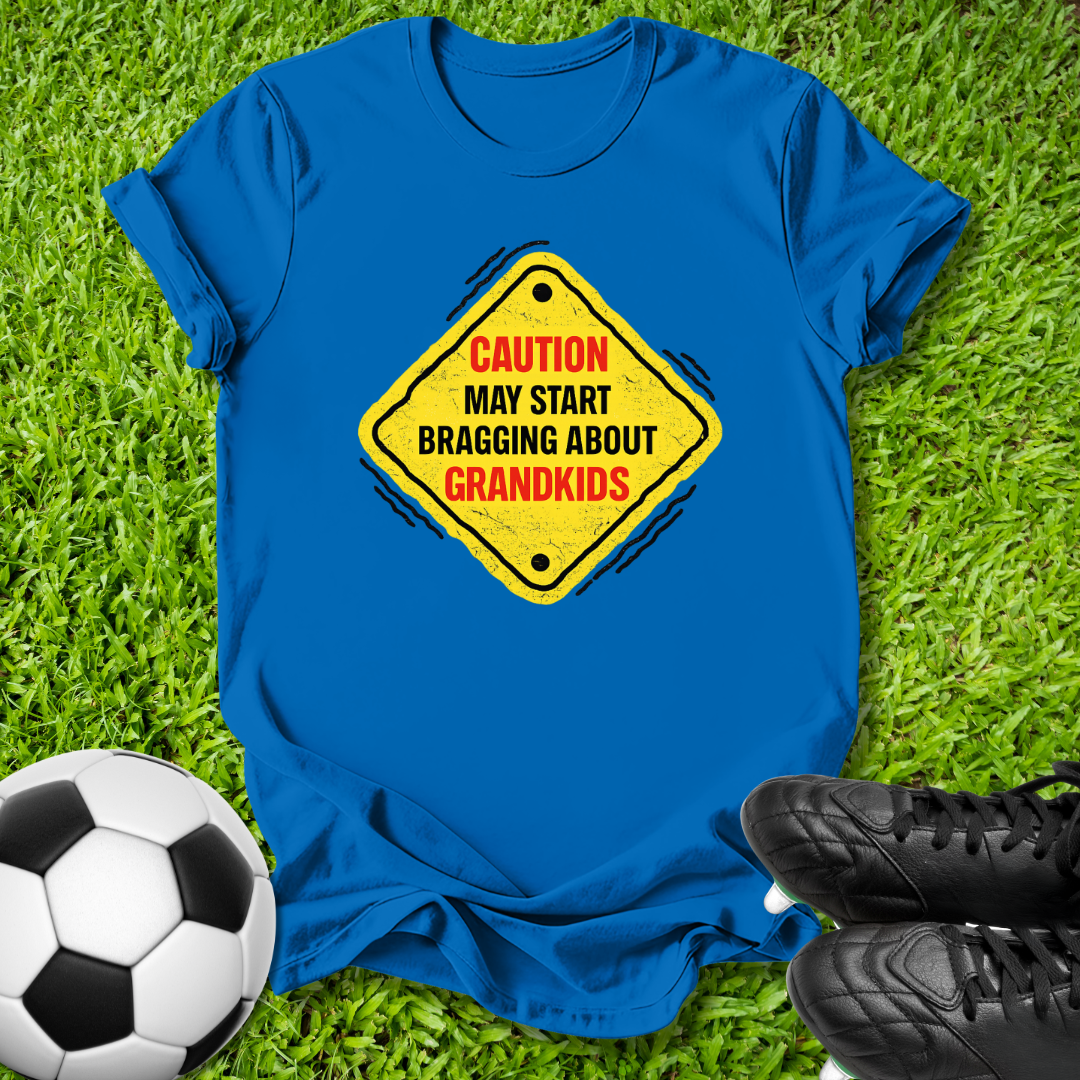 A royal blue short-sleeve t-shirt laid flat on green grass, featuring a yellow diamond-shaped road sign graphic with the text 'CAUTION MAY START BRAGGING ABOUT GRANDKIDS' in red and black. A soccer ball is visible on the left, and black cleats are visible on the right, creating a soccer-themed flat-lay composition.