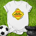 A white short-sleeve T-shirt laid flat on green grass, featuring a yellow diamond-shaped warning sign graphic with the text 'CAUTION MAY START BRAGGING ABOUT GRANDKIDS' in red and black. A soccer ball is at the lower left and black cleats are at the lower right, creating a casual outdoor-style flat lay.