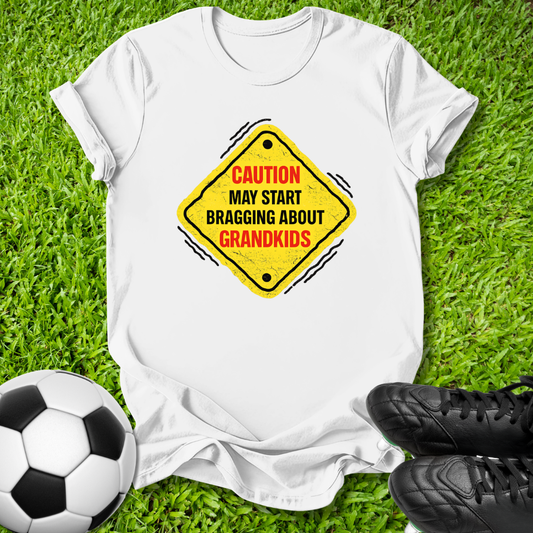 A white short-sleeve T-shirt laid flat on green grass, featuring a yellow diamond-shaped warning sign graphic with the text 'CAUTION MAY START BRAGGING ABOUT GRANDKIDS' in red and black. A soccer ball is at the lower left and black cleats are at the lower right, creating a casual outdoor-style flat lay.