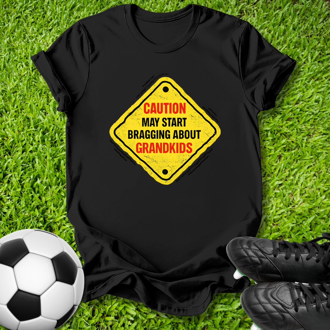 A black short-sleeve t-shirt laid flat on green grass, featuring a yellow diamond-shaped warning sign graphic with red and black text that reads 'CAUTION MAY START BRAGGING ABOUT GRANDKIDS'. A soccer ball is visible at the bottom left and black cleats are at the bottom right.