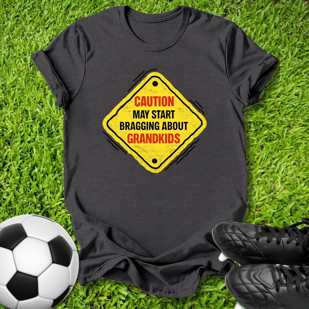 A black short-sleeve t-shirt laid flat on green grass, featuring a yellow diamond-shaped road sign graphic with the text 'CAUTION MAY START BRAGGING ABOUT GRANDKIDS' in red and black. A soccer ball is visible on the left and black cleats are on the right.