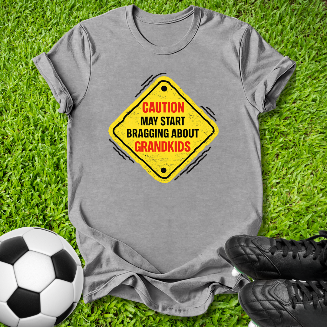 A heather gray t-shirt laid flat on green grass, featuring a yellow diamond-shaped road sign graphic with the text 'CAUTION MAY START BRAGGING ABOUT GRANDKIDS' in red and black. A soccer ball is visible on the bottom left and black cleats are on the bottom right.