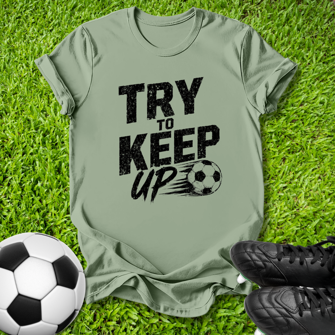 An olive green t-shirt with a black graphic reading 'TRY TO KEEP UP' next to a soccer ball and black cleats, laid flat on green grass.