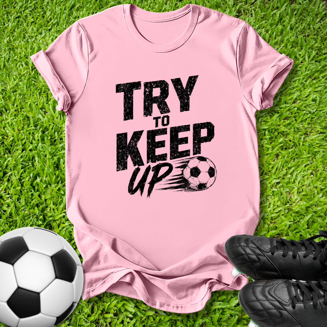 A pink short-sleeve t-shirt with a black graphic reading 'TRY TO KEEP UP' and a soccer ball illustration, laid flat on green grass with a soccer ball and black cleats nearby.