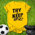 A bright yellow short-sleeve t-shirt with a black distressed graphic reading 'TRY TO KEEP UP' and a soccer ball motif, laid flat on green grass with a soccer ball and black cleats nearby.