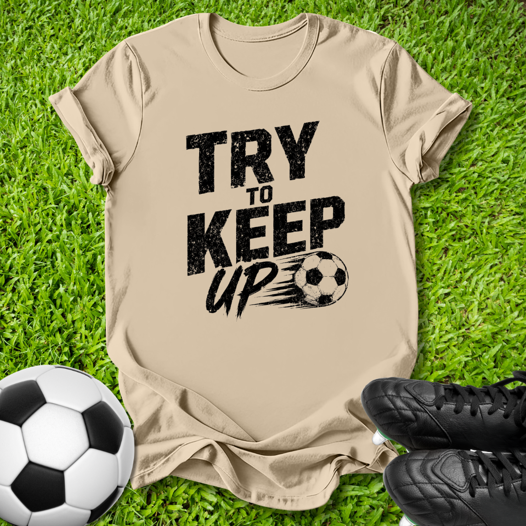 A beige graphic t-shirt laid flat on green grass, featuring the black text 'TRY TO KEEP UP' next to a soccer ball and black cleats.