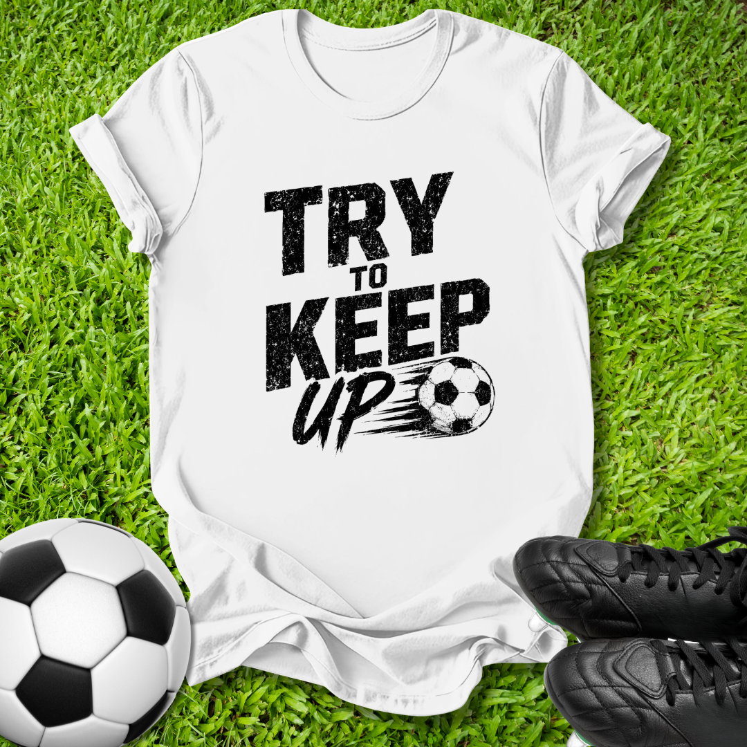 A white short-sleeve graphic T-shirt laid flat on green grass, with black text reading 'TRY TO KEEP UP' and a soccer ball illustration. A soccer ball and black cleats are placed around it as props.