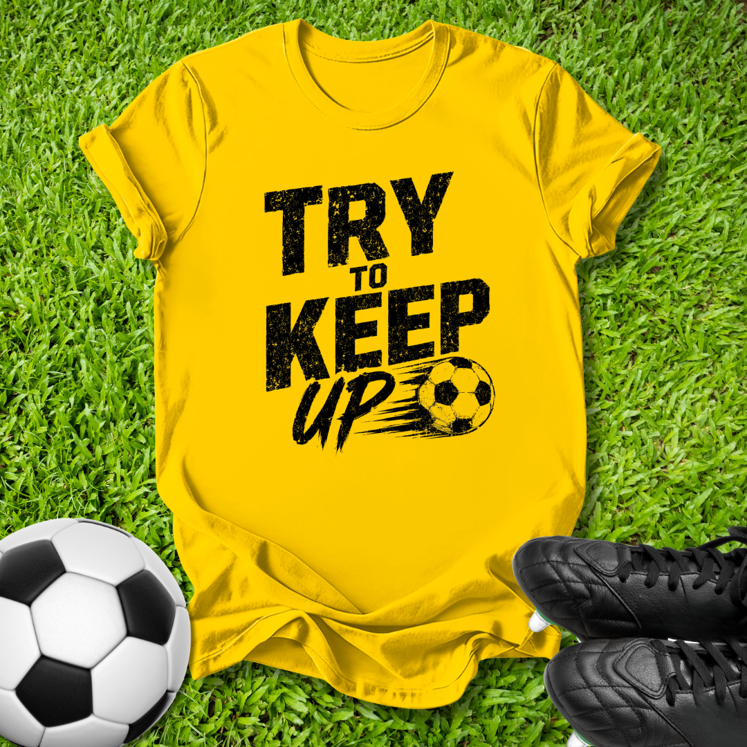 A bright yellow short-sleeve t-shirt with a black distressed graphic reading 'TRY TO KEEP UP' and a soccer ball motif, laid flat on green grass with a soccer ball and black cleats nearby.