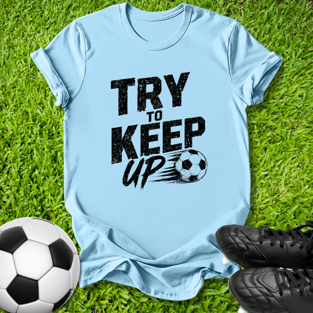 A light blue t-shirt with a black graphic reading 'TRY TO KEEP UP' and a soccer ball illustration, laid flat on green grass with a soccer ball and black cleats nearby.