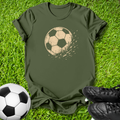 An olive green short-sleeve t-shirt with a distressed beige soccer ball graphic on the chest, laid flat on green grass with a soccer ball and black cleats as props.