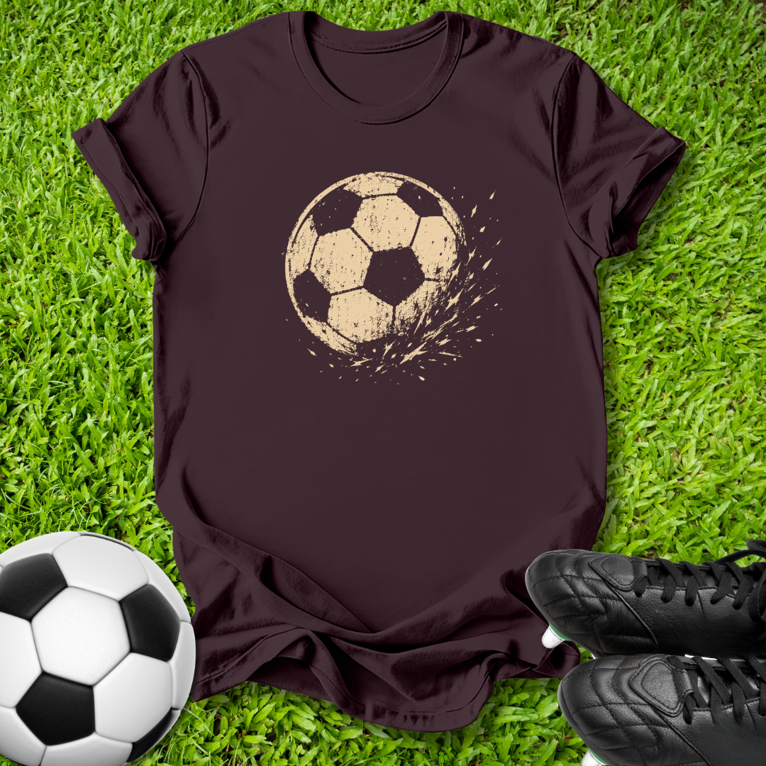 A dark brown short-sleeve t-shirt with a distressed beige soccer ball graphic on the chest, laid flat on green grass with a soccer ball and black cleats as props.