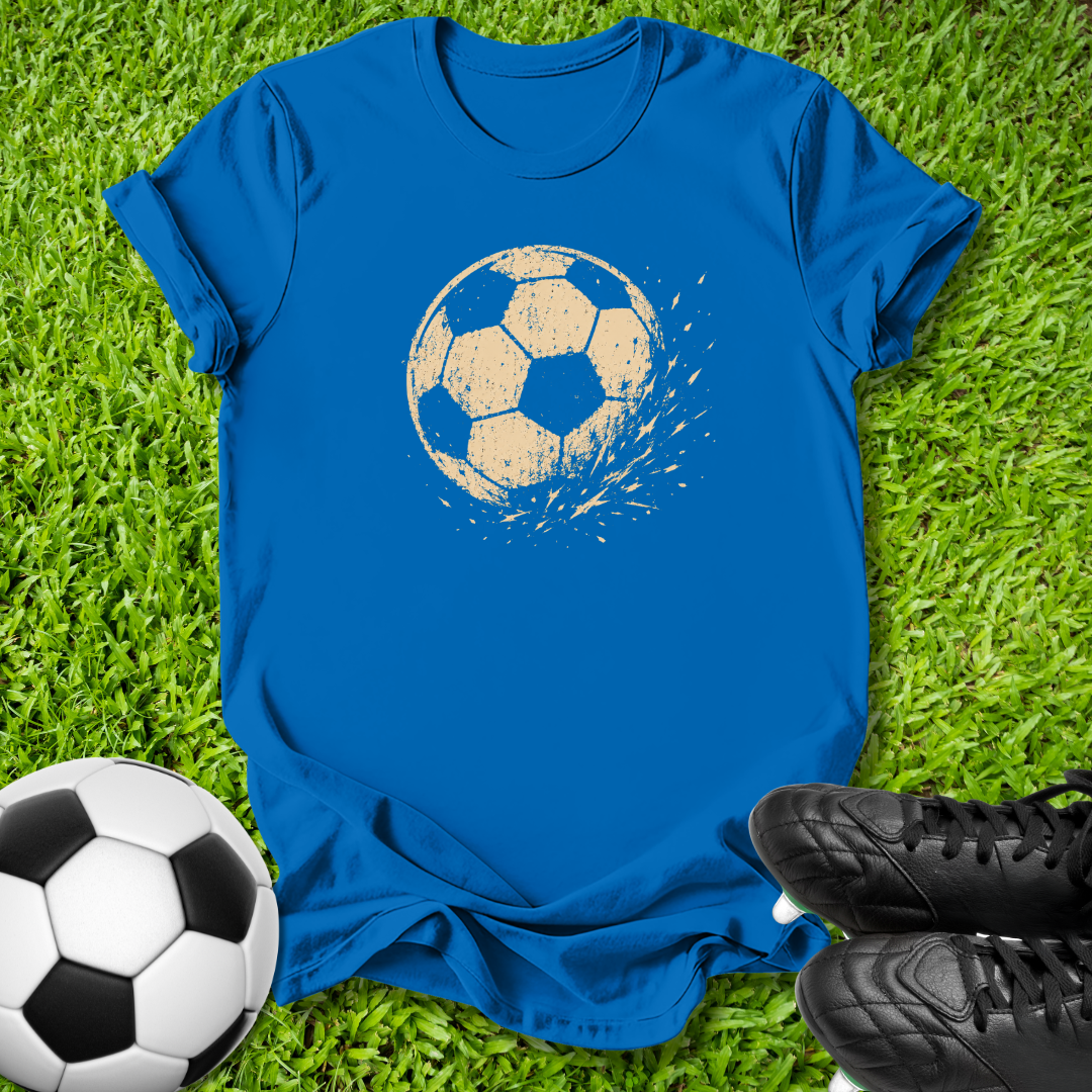 A royal blue short-sleeve t-shirt laid flat on green grass, featuring a distressed graphic of a soccer ball in the center. A real soccer ball is visible in the bottom-left corner, and black cleats are visible in the bottom-right corner, creating a sports-themed flat lay.