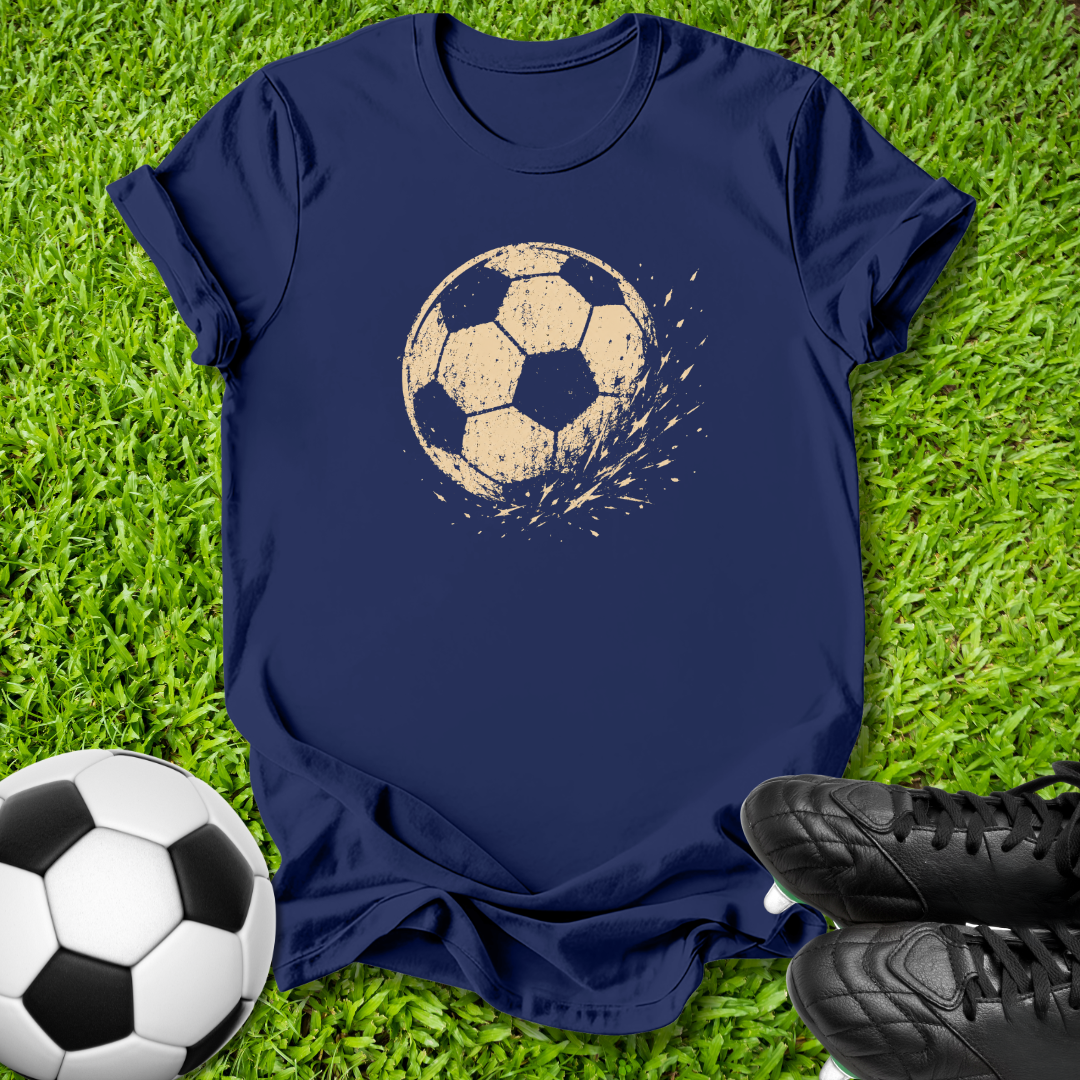 A navy blue short-sleeve t-shirt with a distressed beige soccer ball graphic on the chest, laid flat on green grass with a soccer ball and black cleats nearby.