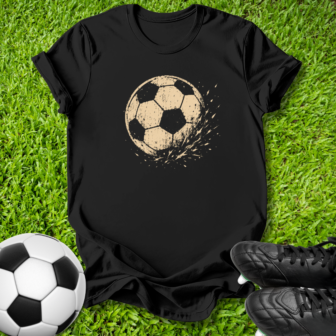 A black short-sleeve t-shirt laid flat on green grass, featuring a distressed beige soccer ball graphic on the chest. A real soccer ball and black cleats are placed around it as props.