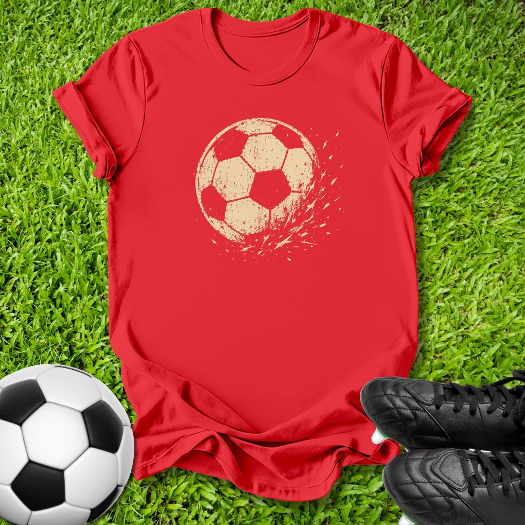 A red short-sleeve t-shirt with a distressed beige soccer ball graphic on the chest, laid flat on green grass with a soccer ball and black cleats as props.