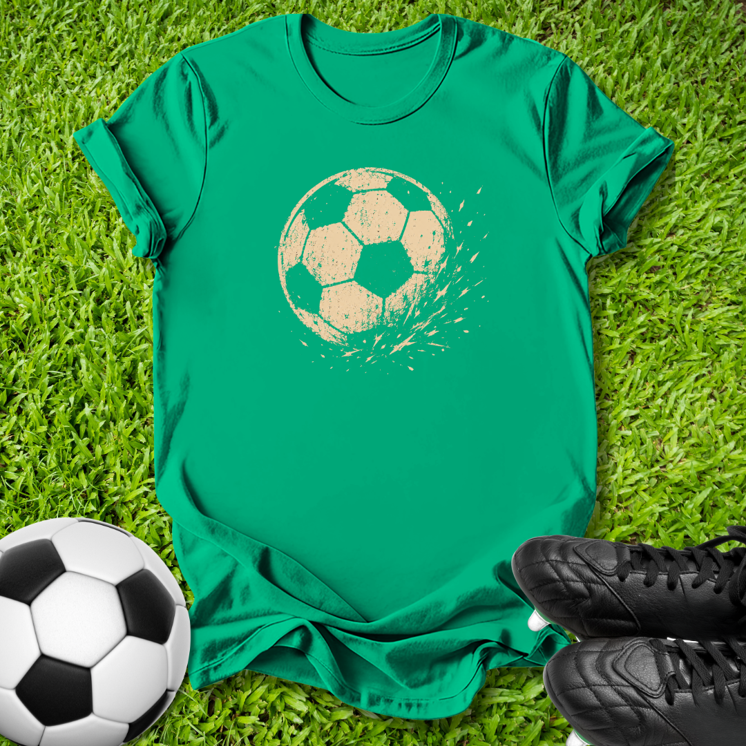 A green short-sleeve T-shirt laid flat on grass, featuring a distressed beige soccer ball graphic on the chest. A black-and-white soccer ball sits at the lower left, and black cleats are at the lower right, creating a soccer-themed flat lay.