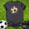 A gray short-sleeve t-shirt laid flat on green grass, featuring a distressed tan soccer ball graphic on the chest. A real soccer ball is placed at the bottom left, and black cleats are at the bottom right, creating a soccer-themed flat lay.
