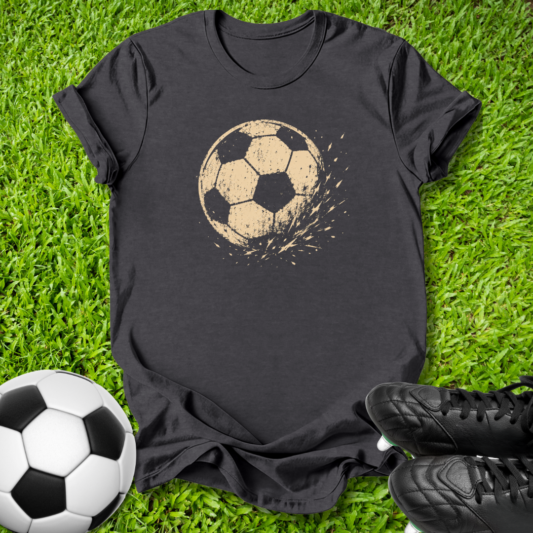 A gray short-sleeve t-shirt laid flat on green grass, featuring a distressed tan soccer ball graphic on the chest. A real soccer ball is placed at the bottom left, and black cleats are at the bottom right, creating a soccer-themed flat lay.
