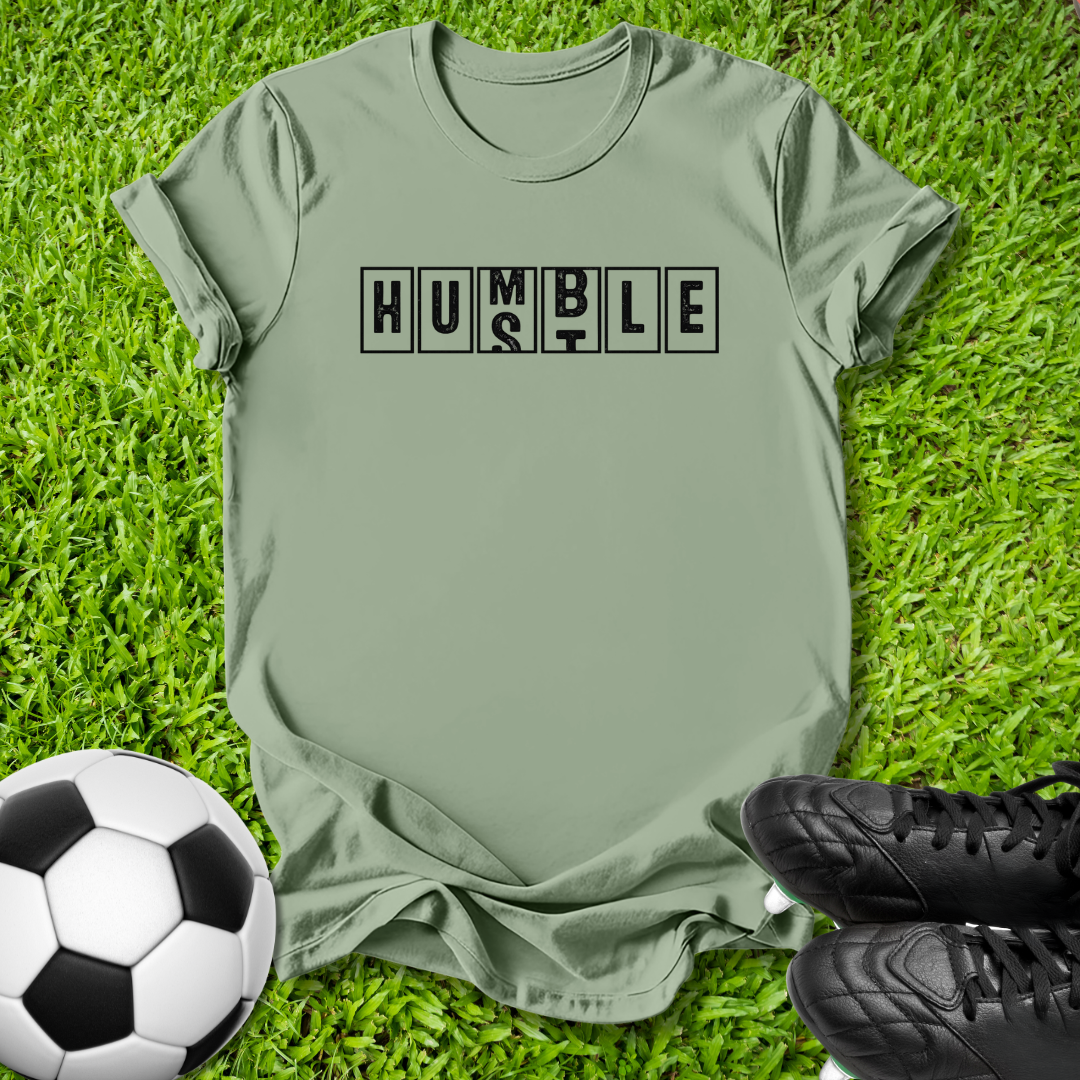 An olive green short-sleeve t-shirt with the word 'HUMBLE' printed in black block letters across the chest, laid flat on green grass with a soccer ball and black cleats as props.