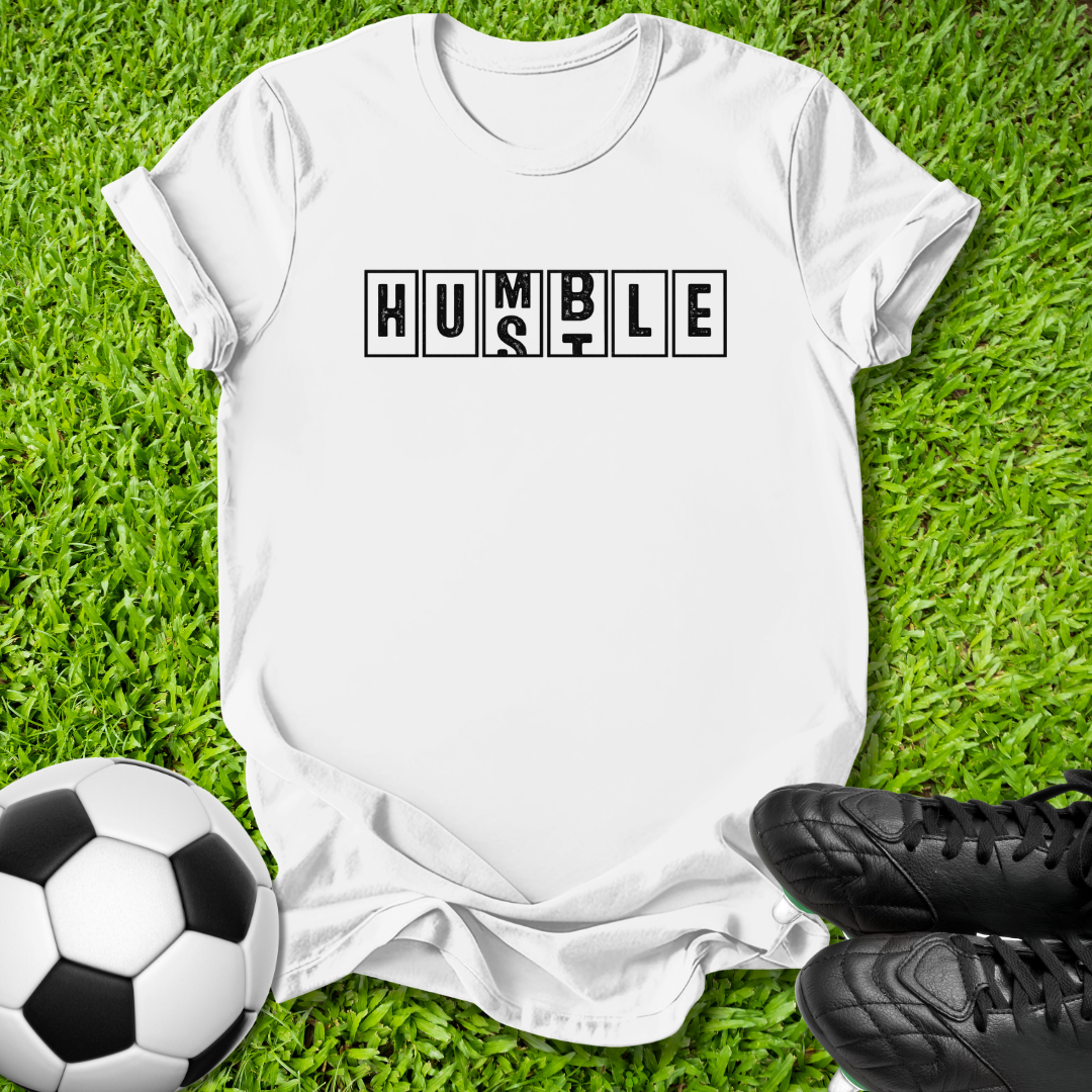 A white short-sleeve T-shirt laid flat on green grass, with the word 'HUMBLE' printed across the chest in black block letters. A soccer ball appears at the lower left, and black cleats are at the lower right, creating a styled outdoor flat-lay.