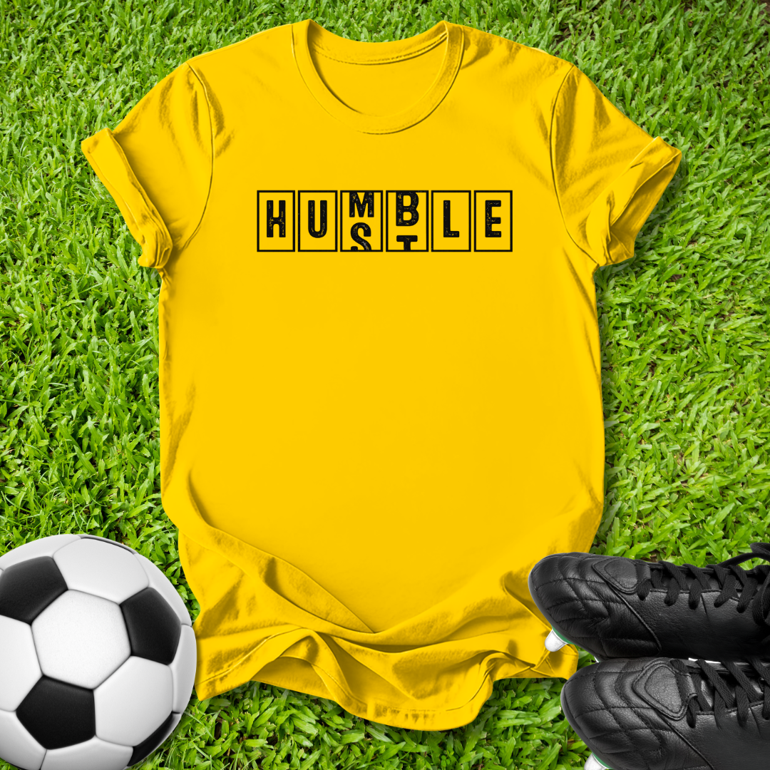 A bright yellow t-shirt with the black text 'SOMEWHERE ON THE SIDELINES' printed across the chest, laid flat on green grass with a soccer ball and black cleats as props.