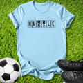 A light blue short-sleeve t-shirt laid flat on green grass, featuring the word 'HUMBLE' printed across the chest in black block letters. A soccer ball is visible in the lower-left corner and black cleats are in the lower-right corner.