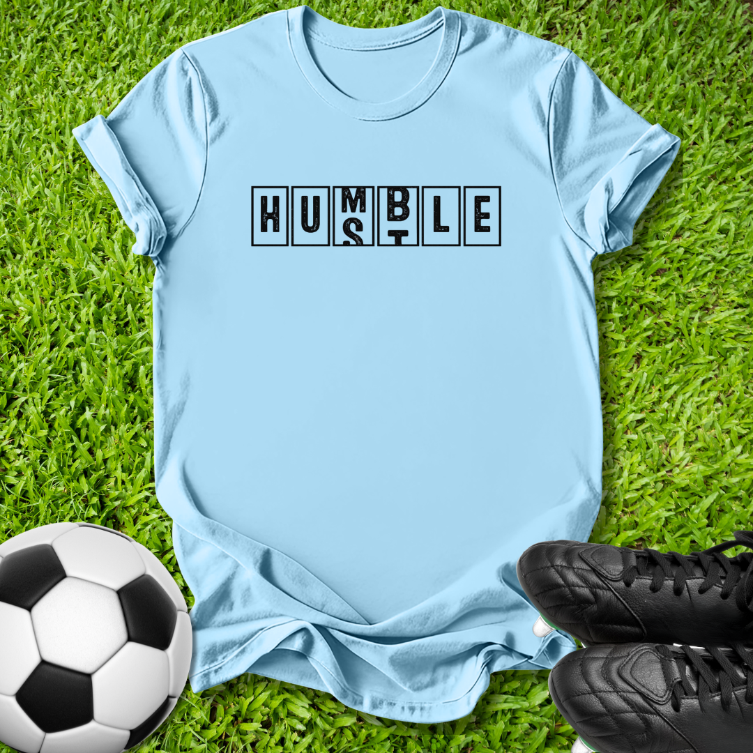 A light blue short-sleeve t-shirt laid flat on green grass, featuring the word 'HUMBLE' printed across the chest in black block letters. A soccer ball is visible in the lower-left corner and black cleats are in the lower-right corner.