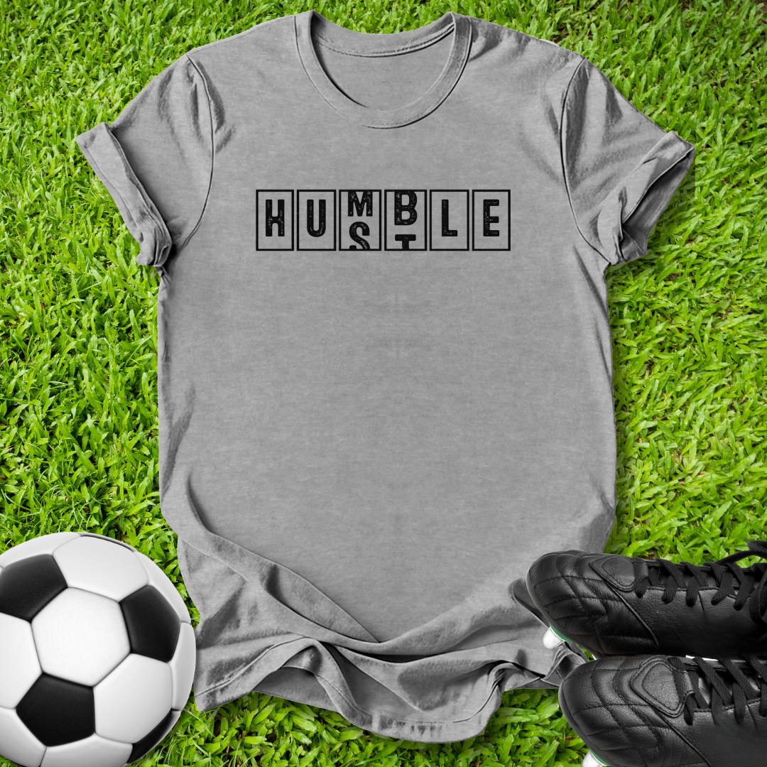 A heather gray short-sleeve t-shirt with the word 'HUMBLE' printed across the chest in black block letters, laid flat on green grass with a soccer ball and black cleats nearby.