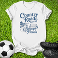 A white short-sleeve graphic T-shirt laid flat on green grass, with a blue printed slogan reading 'Country Roads Lead to Soccer Fields' and a small soccer illustration. A soccer ball appears at the lower left and black cleats at the lower right, creating a styled outdoor flat-lay.