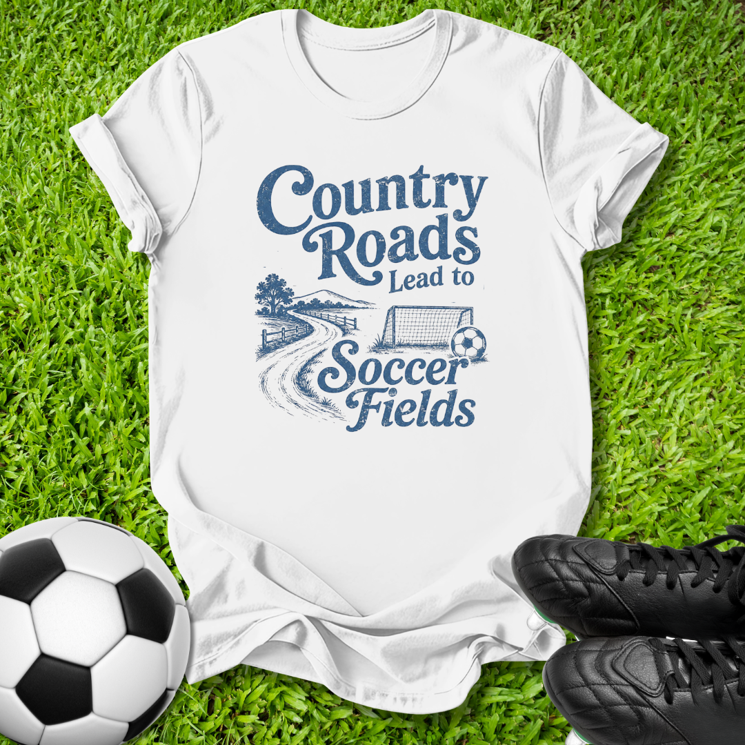 A white short-sleeve graphic T-shirt laid flat on green grass, with a blue printed slogan reading 'Country Roads Lead to Soccer Fields' and a small soccer illustration. A soccer ball appears at the lower left and black cleats at the lower right, creating a styled outdoor flat-lay.