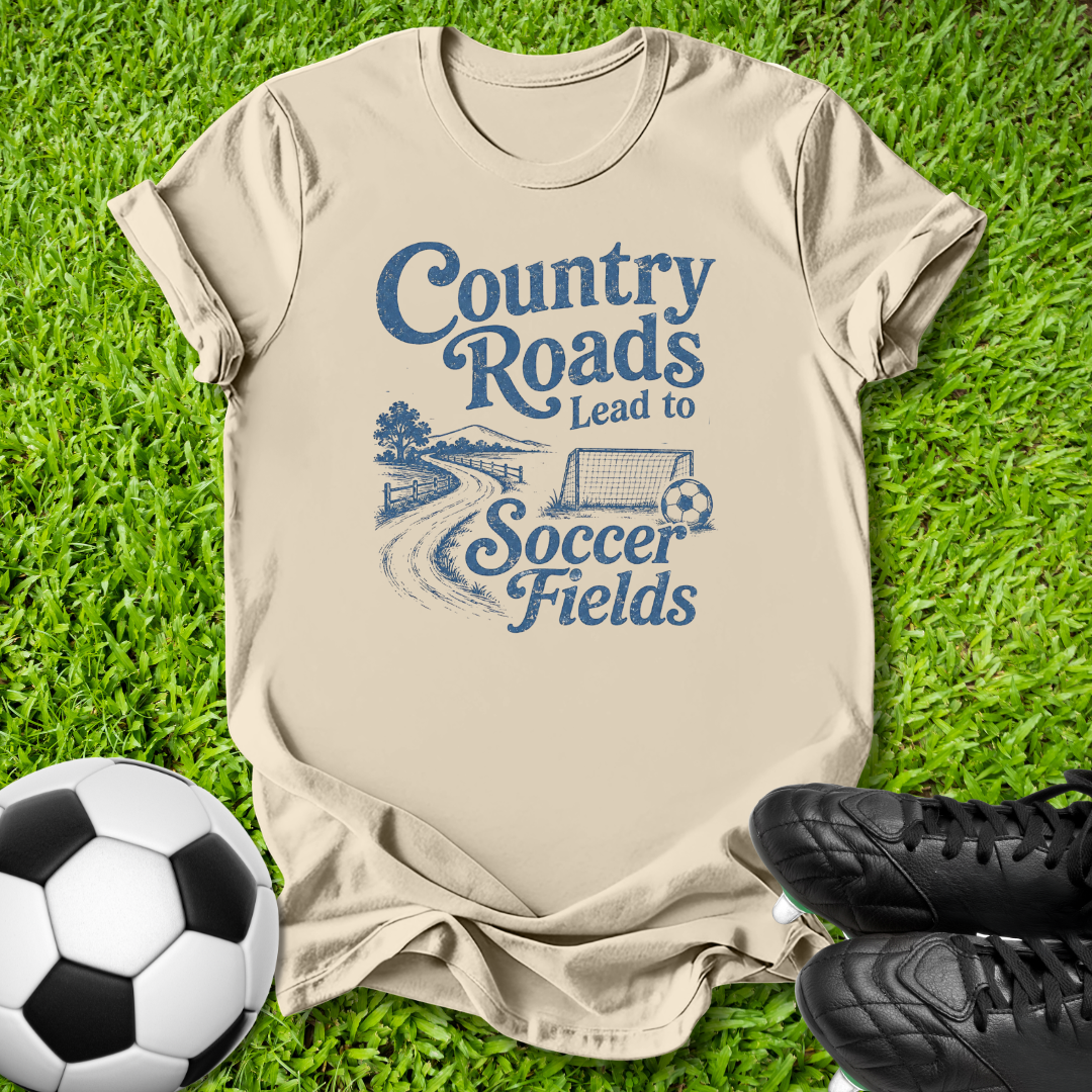 A beige graphic t-shirt laid flat on green grass, featuring a blue printed design that reads 'Country Roads Lead to Soccer Fields' with a stylized road, tree, and soccer ball graphic. A soccer ball and black cleats are placed around it as props.