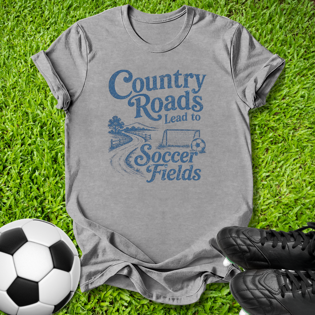 A heather gray t-shirt with a blue graphic reading 'Country Roads Lead to Soccer Fields' is laid flat on green grass, styled with a soccer ball on the left and black cleats on the right.