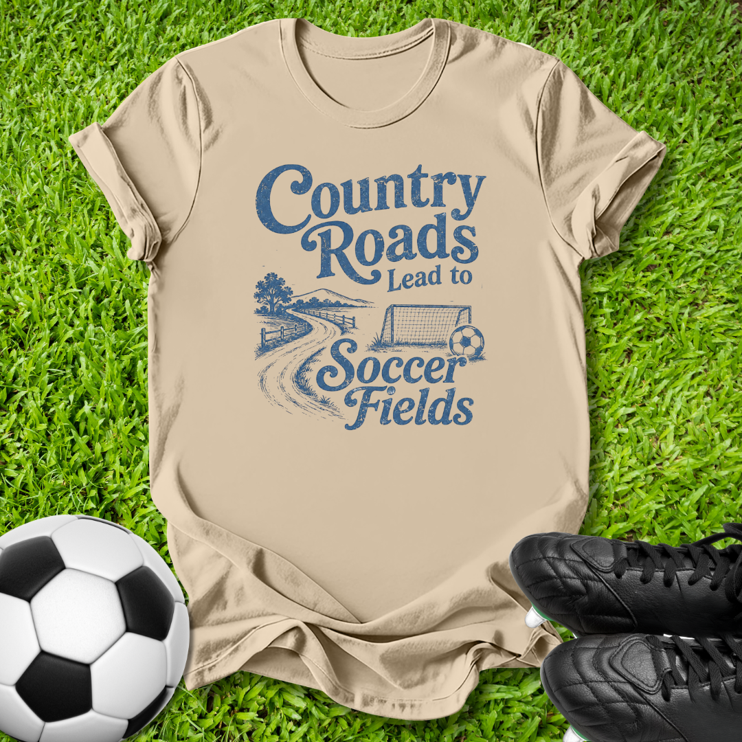 A beige short-sleeve t-shirt with a blue graphic reading 'Country Roads Lead to Soccer Fields' laid flat on green grass, styled with a soccer ball and black cleats.